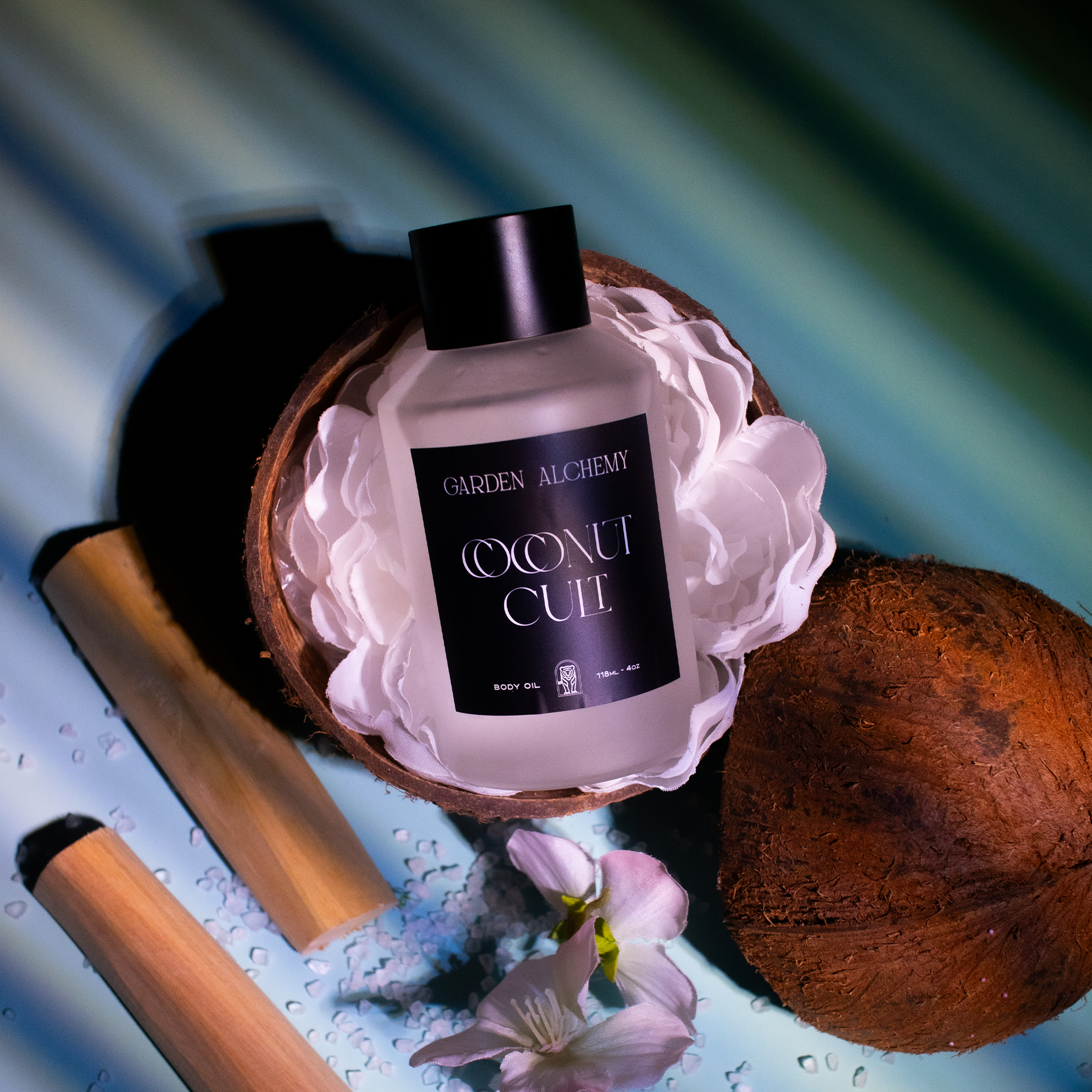 A bottle of GARDEN ALCHEMY COCONUT CULT PERFUME BODY OIL rests in a halved coconut among white petals, with bamboo sticks, flower petals, and blue bath salts placed nearby on a striped surface.