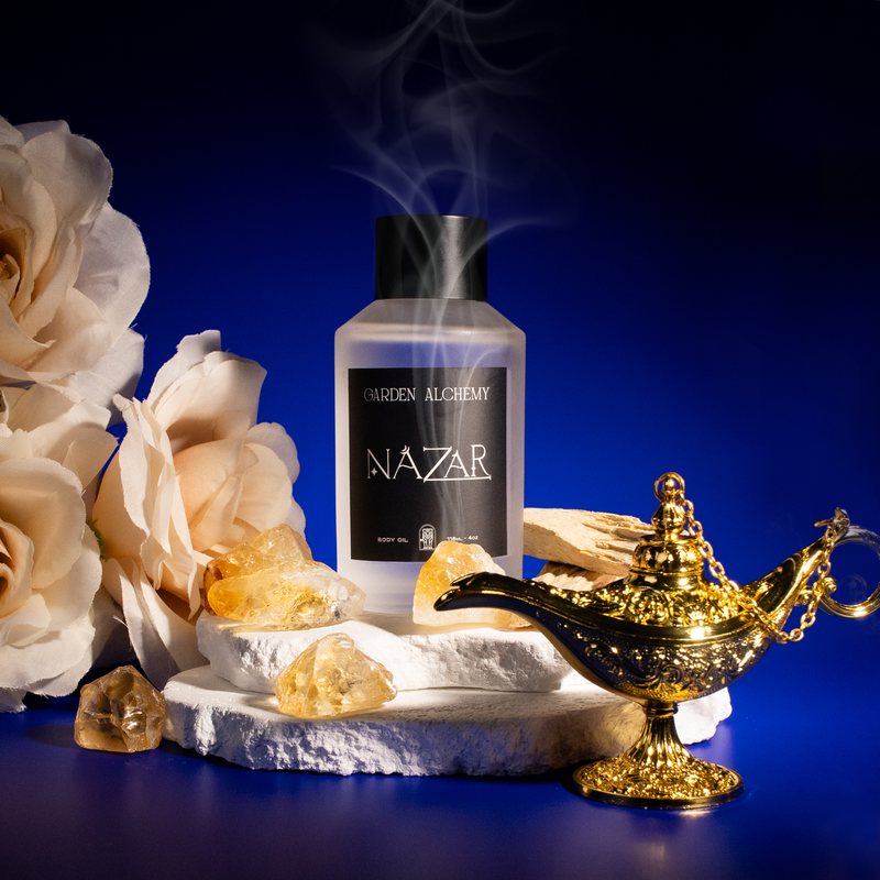 A GARDEN ALCHEMY NAZAR BODY OIL bottle rests on white stones with crystals, pale roses, and a golden genie lamp, evoking the mystical aura of the evil eye against a blue smoke-filled background.